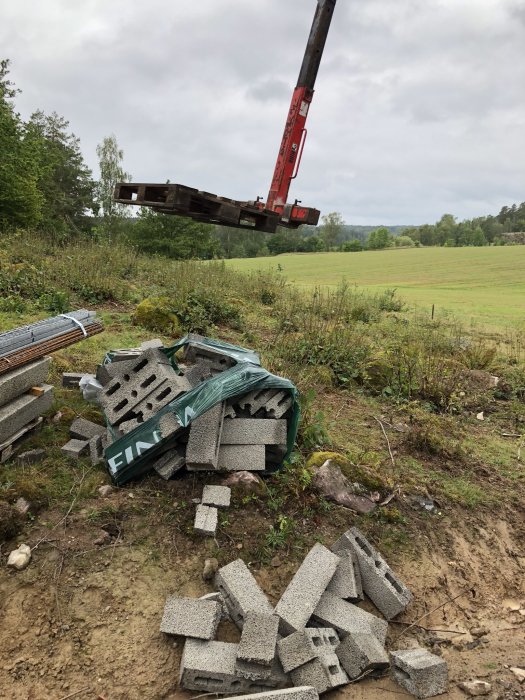 En byggarbetsplats med armeringsjärn och staplade betongblock, några spillda på marken, med en kran som lyfter material i bakgrunden.