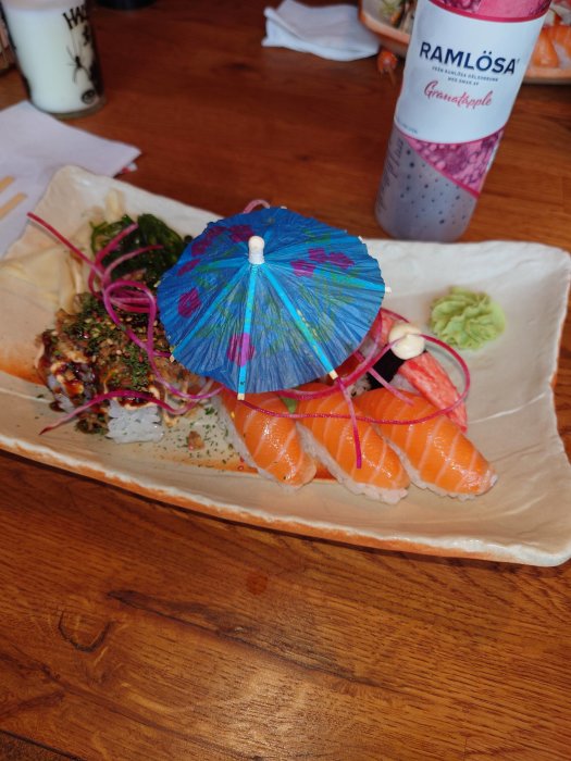 Läckert upplagd sushi med cocktailparaply och granatäpple Ramlosa-flaska på ett träbord.