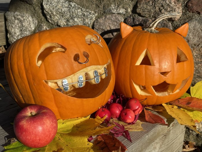 Två utsmyckade pumpor, en med tandställning och en med piercing, bredvid ett äpple och löv.