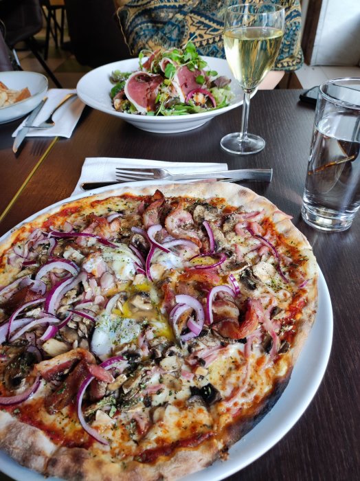 Pizza med lök, svamp och bacon på bord med sallad och glas vin.