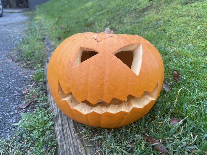 Utskuren Halloween-pumpa med ansikte placerad på gräs vid en träkant.