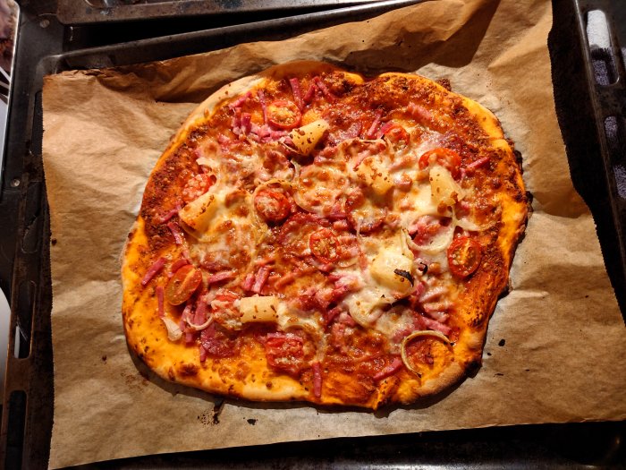 Hembakad pizza med tomatsås, ost, skinka, ananas och tomat på bakplåtspapper.