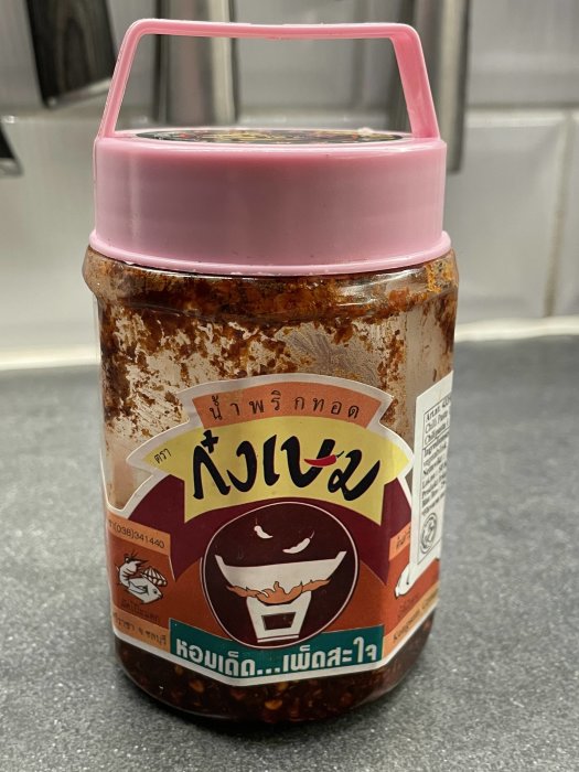 En öppnad burk med stark chilipasta, med text på thailändska, står på en köksbänk.