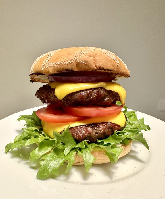 Hamburgare med två biffar, ost, tomat, sallad och bröd på vit tallrik.