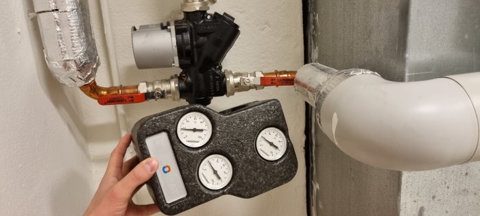 Hand håller en mätutrustning med tryckmätare framför en värmesysteminstallation.