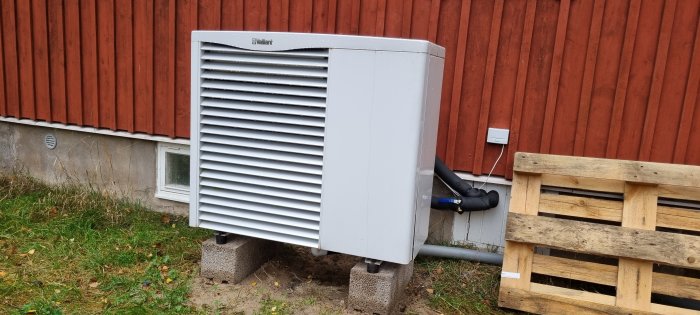 Luftvärmevärmepump placerad utomhus framför röd trävägg med synliga in- och utloppsrör.