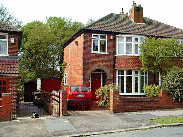 Rödtegelfasad halvfristående hus med sluttande tak och parkerad bil framför.