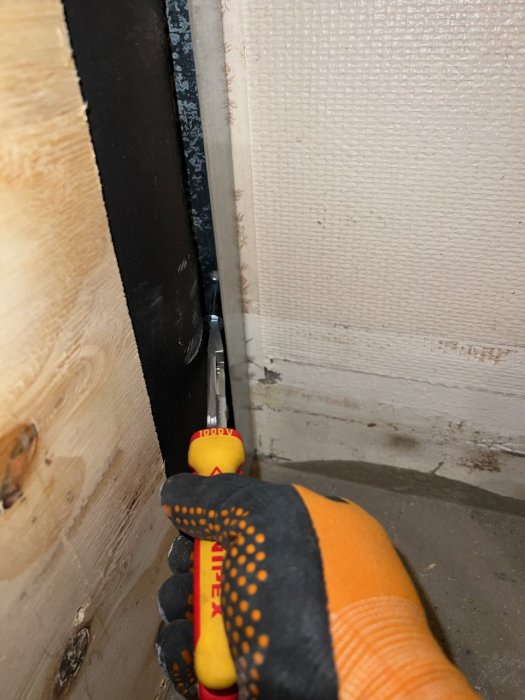 Närbild på en hand i skyddshandskar som skär igenom isolering med en kniv nära en vägg med plywood och ångspärr.
