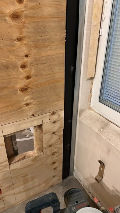 Delvis konstruerad vägg med plywood och synliga isoleringsmaterial vid hörn av rum, nära fönster och verktyg.