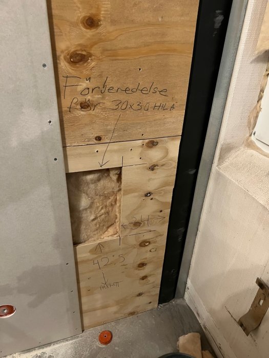 Renoveringsdetalj med isolering, plywood och märkningar för inspektionslucka i en schaktvägg.