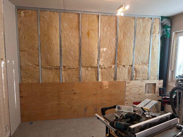 Isolering och delvis monterad plywood på en inomhusvägg under renovering, med verktyg i förgrunden.