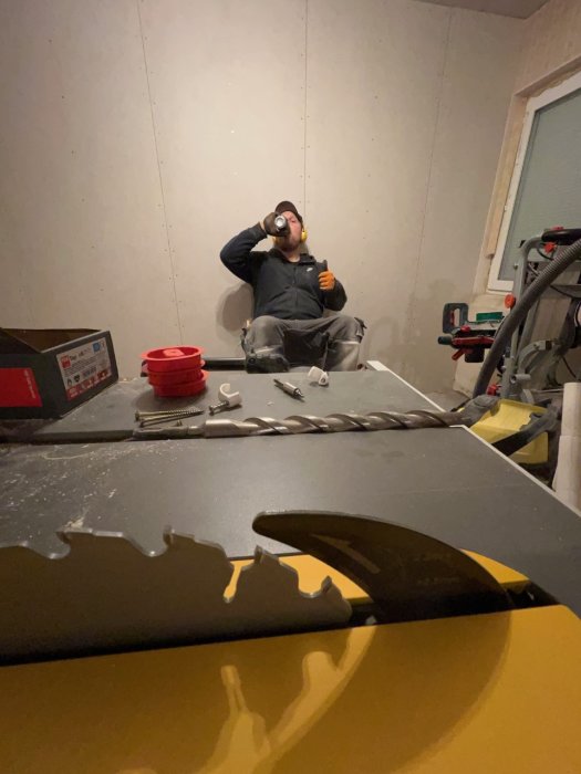 Person i arbetskläder tar en paus med en öl i halvfärdigt renoverat rum med byggmaterial.