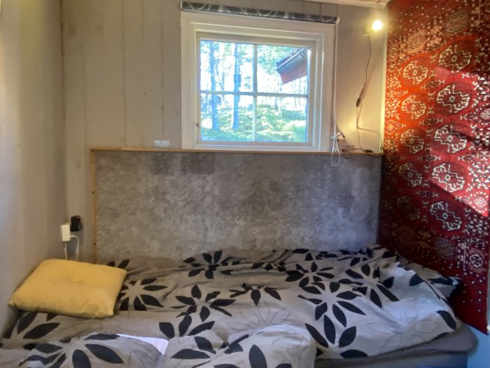 Sovrum med en grå matta på väggen, säng med mönstrat överkast och röd vävnad på sidan.