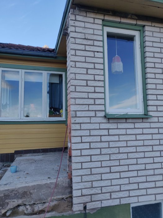 Husfasad med vit tegelimitation, fönster med röd lampa inuti och delvis rivet trappsteg.