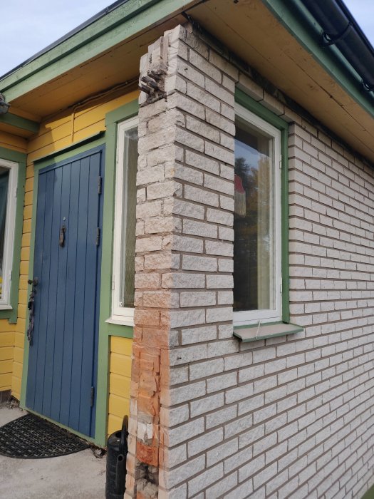Skadad hörndel av en tegelstenfasad vid en husentré visar tecken på förfall och behov av reparation.