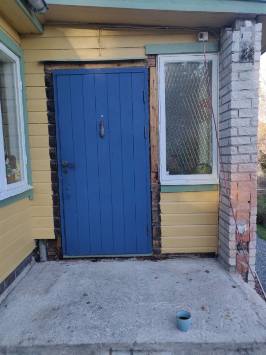 Blå ytterdörr på hus med skadat träverk och en järnkedja hängandes, stenbelagd trappa framför.