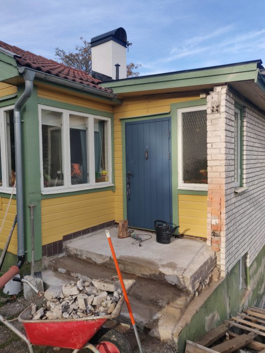 Husfasad under renovering med avlägsnad trappa och samlade betongbitar i en röd skottkärra.