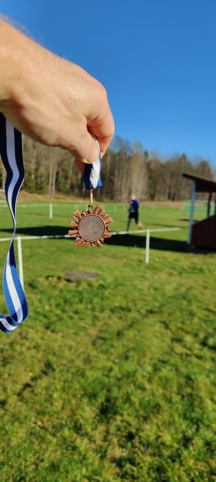 Hand håller upp en bronsmedalj med band, bakgrunden visar en person som joggar på en idrottsplats.