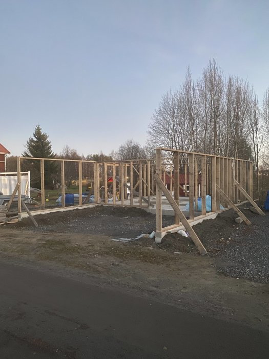 Nybyggda trästommar för en carport på grusunderlag i ett bostadsområde under skymningen.