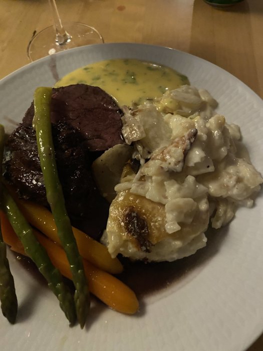 En middagstallrik med oxfilé, potatisgratäng och ångade gröna sparris samt morötter, serverad med sås.