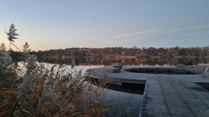 Sjövy vid solnedgång med en person som sitter på en cirkulär träbrygga omgiven av vass.