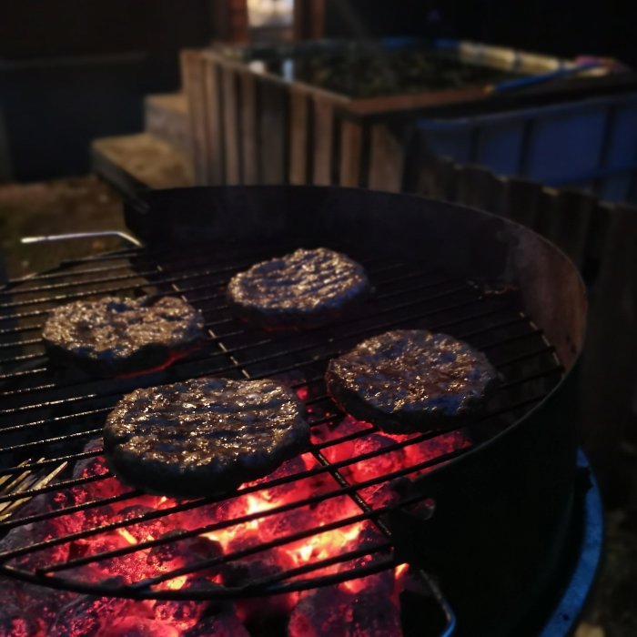 Hamburgare av högrevsfärs grillas över glödande kol på en utomhusgrill på kvällen.