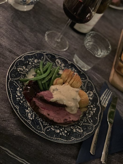 Älgstek med hasselbackspotatis, kantarellsås och haricots verts på mönstrad tallrik, vinglas i bakgrunden.