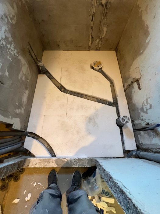 Vy ovanifrån av golvvärmeinstallation med slangar, spårad cellplast och verktyg i ett orenoverat badrum.