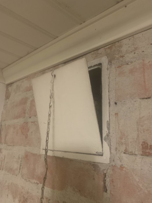 Vit lucka på kedja framför öppning i tegelvägg, installationslucka för ventilation.