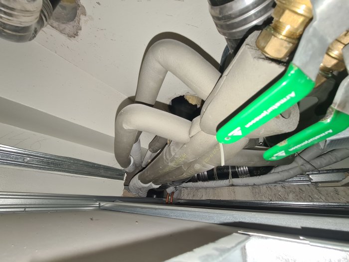 Vy underifrån av rörledningar och ventiler i ett tak, isolerade och märkta med gröna handtag.