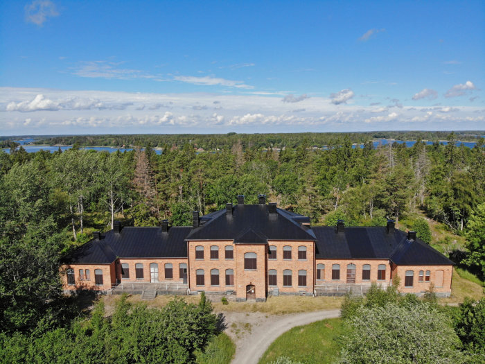 Flygbild över historiskt tegelhus med omgivande skog och havsutsikt under klar himmel.