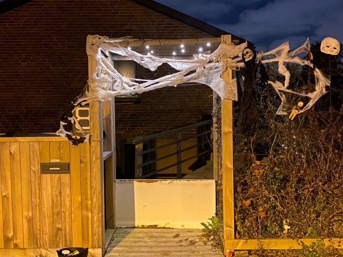 Trädgård entré dekorerad med Halloween-tema: spindelväv, skelett, spöken, kvällsbelysning mot tegelbakgrund.