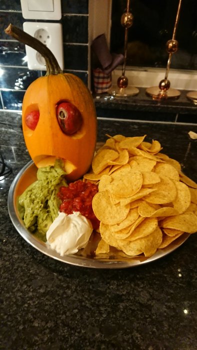 En pumpa med ansikte, guacamole, salsa, gräddfil och nachos på serveringsfat. Festligt tilltugg med Halloween-tema.