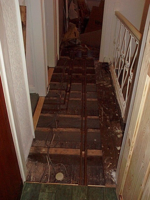 Ett uppbrutet golv i en korridor med synliga golvbjälkar och bråte. Renovering eller skadeåtgärd pågår.