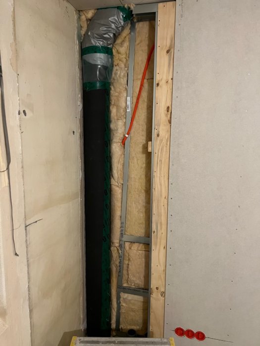 Delvis konstruerad vägg, isolering, ventilationsslang, röda plastlock på golvet, byggarbetsplats.
