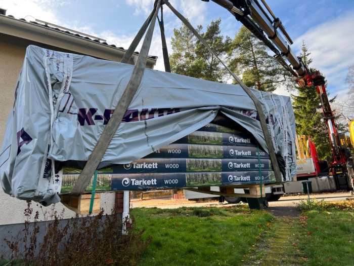 Lastbil med kran lyfter stora paket av Tarkett-golv. Bostadsområde, dagtid, byggmaterial, utomhus.