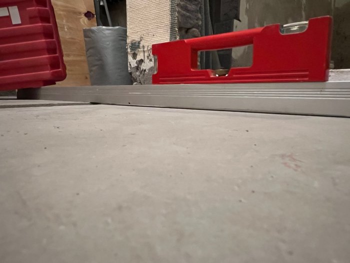 Rött verktyg, troligtvis vattenpass, på betonggolv i arbetsmiljö med röd låda och byggmaterial i bakgrunden.