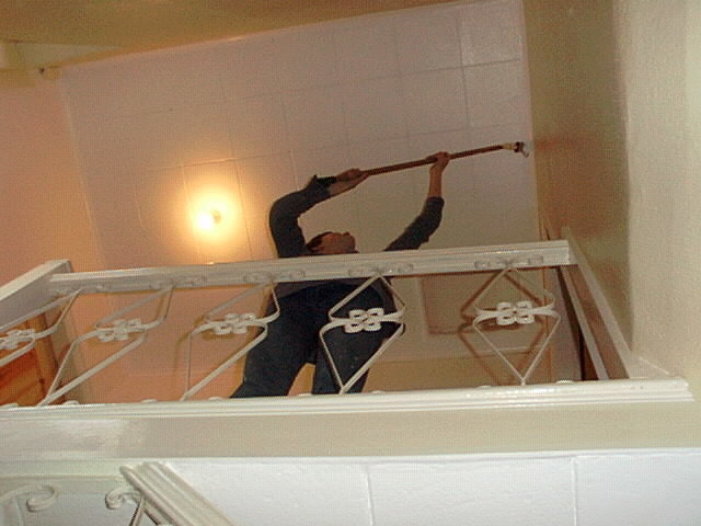 Person målar tak, använder förlängningsskaft, inomhus, trapphus, vit vägg, säkerhetsräcke, arbetskläder.