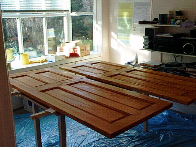 Ett upplyst arbetsrum med nybehandlade trädörrar på ett bord, skyddsplast på golvet, bakgrundsfönster och lådor.