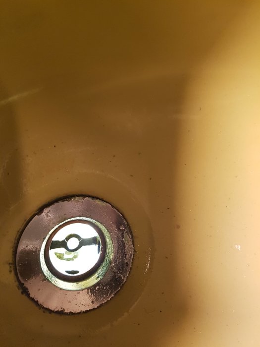 Närbild på en sliten handfatavlopp med metallring och speglande vatten, gulaktig bakgrundston.