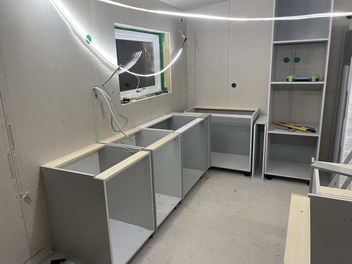 Ett pågående köksinstallation med grå skåpstommar, verktyg, och LED-ljusslinga i ett underkonstruerat rum.
