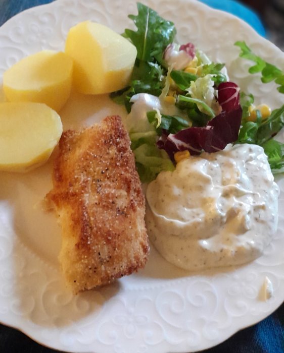 En tallrik mat innehållande panerad fisk, kokta potatisar, sallad och remouladsås.