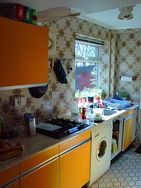 Ett retrokök med blommigt tapetmönster, orange skåp, tvättmaskin och rörigt diskbänk.