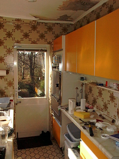 Gammalt kök med orange skåp och blommigt tapet, oordning, behöver renovering, dagsljus, utsikt till naturen.