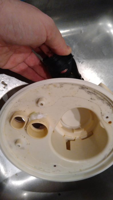 En hand håller en smutsig del av en vit plastapparat, troligtvis från en köksmaskin, över ett diskho.