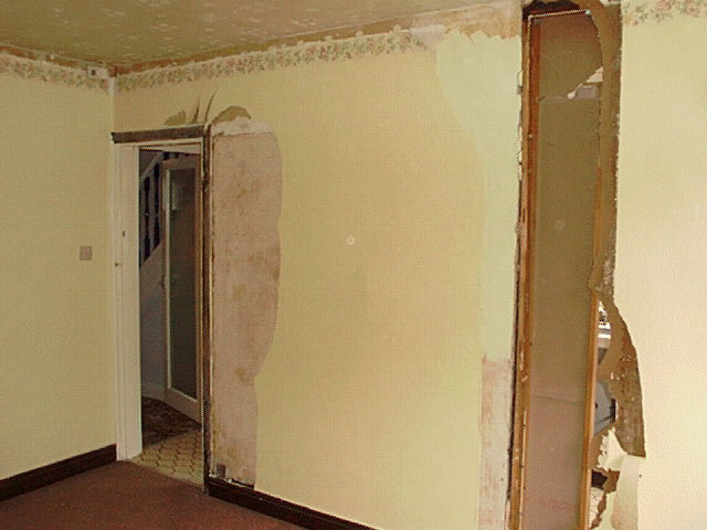 Korridor i hus med avskalad tapet, skadad vägg och dörrpost, behövande renovering eller reparation.