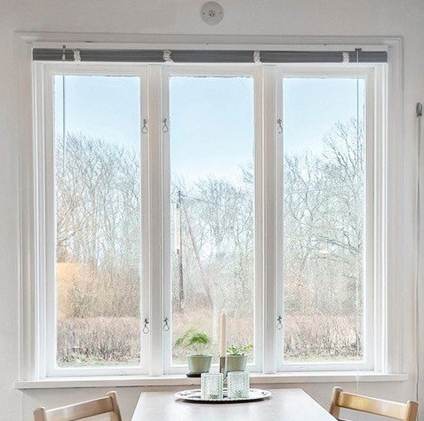 Ljust rum. Stort fönster med utsikt över natur. Bord med dekorationer. Vita gardiner. Trästolar. Dagtid.