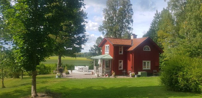 Rött svenskt hus med vita knutar, grönskande trädgård, soligt, utemöbler, lantlig idyll, harmoniskt.