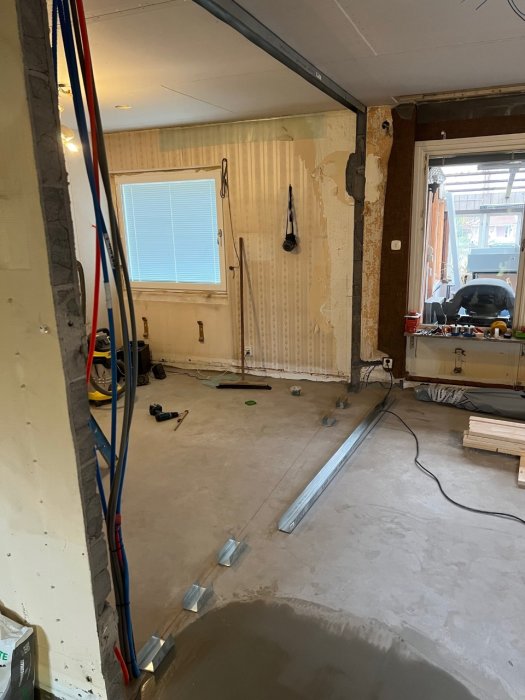 Renovering pågår, rör och verktyg synliga, avskalade väggar, ojämnt golv, fönster, delvis strippat tak, byggmaterial.