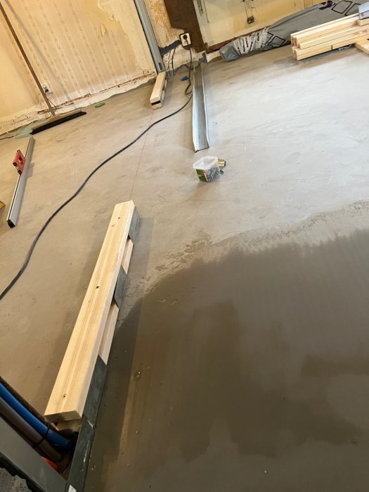Inomhusbyggarbetsplats med färskt gjutet golv, verktyg, trämaterial och en elektrisk kabel.